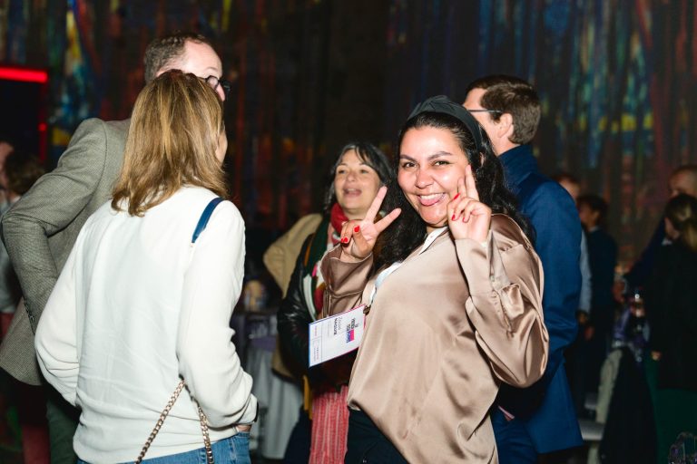 Une femme en chemisier satiné, souriante et faisant le signe de la paix avec les deux mains, se tient parmi quatre autres personnes en conversation lors de la French Tech Night 2024. Elle a un cordon autour du cou, et l'arrière-plan est faiblement éclairé par des lumières colorées et des figures floues.