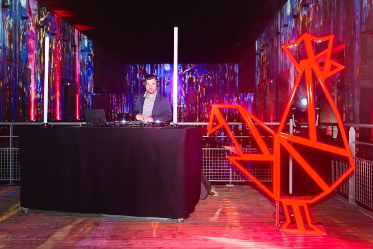 Un DJ se tient derrière une table drapée de noir avec du matériel de mixage dans une salle faiblement éclairée. La salle est dotée d'un éclairage vibrant et abstrait sur les murs. À droite du DJ se trouve une grande sculpture géométrique de coq rouge illuminée, symbolisant la French Tech Night 2024. Un parquet en bois et des balustrades en métal sont visibles.