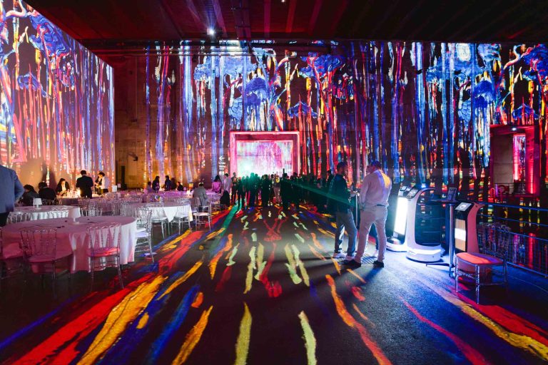 Une exposition d'art à la French Tech Night 2024 avec des projections lumineuses vibrantes et multicolores sur les murs et le sol, mettant en scène des motifs et des figures abstraits. Plusieurs personnes se mêlent et observent l'exposition. À gauche, des tables rondes avec des nappes et des chaises blanches sont installées. Deux kiosques robotisés sont présents à droite.