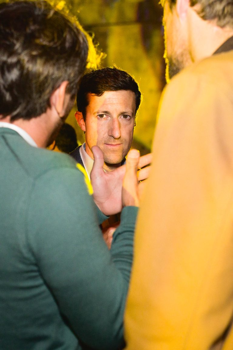 Trois hommes discutent, deux d'entre eux sont vus de dos et un autre est face à la caméra. L'homme face à la caméra a les cheveux courts et foncés et porte un blazer foncé sur une chemise claire. La photo, prise lors de la French Tech Night 2024, est éclairée par une teinte jaunâtre, créant un décor intérieur chaleureux.