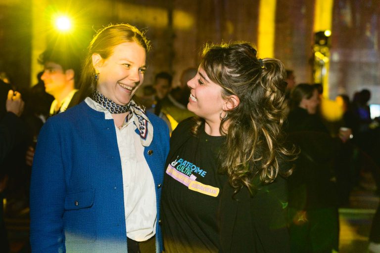 Deux femmes souriantes, l'une en veste bleue et écharpe, l'autre en chemise noire à motifs colorés, discutent lors de la French Tech Night 2024. La salle chaleureusement éclairée est animée et conviviale, remplie de monde. Une lumière jaune ajoute à l'atmosphère chaleureuse tandis qu'elles se regardent.