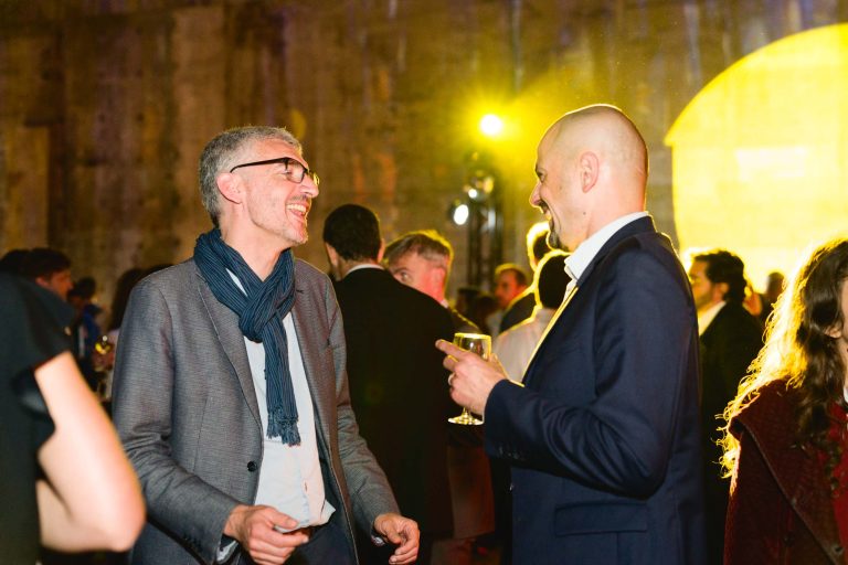 Lors de la French Tech Night 2024, deux hommes en tenue de soirée discutent et rient lors d'un événement en salle, une lumière jaune chaleureuse illuminant la salle. L'homme de gauche porte un costume gris et une écharpe bleue, tandis que l'homme de droite porte un costume bleu marine et tient un verre. D'autres participants sont visibles en arrière-plan.