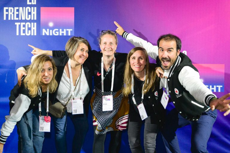 Cinq personnes se tiennent devant un fond violet sur lequel est écrit « LQ FRENCH TECH NIGHT », toutes souriantes et posant avec enthousiasme. Elles portent des cordons de l'événement et des tenues décontractées, certaines portant des vestes noires assorties. Deux personnes aux extrémités pointent leurs mains vers l'extérieur dans un geste vivant, célébrant la French Tech Night 2024.