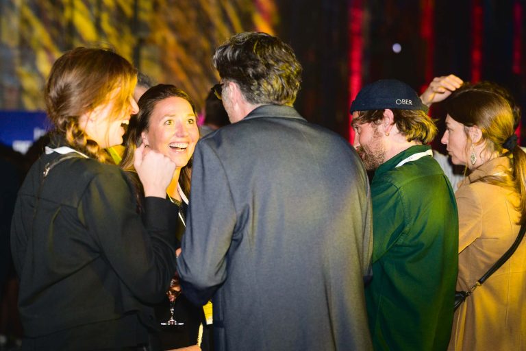 Un groupe de cinq personnes discutent à la French Tech Night 2024. Deux femmes à gauche sourient et discutent. Un homme en costume tourne le dos à la caméra, face au groupe. Un homme en chemise verte avec une casquette sombre et une femme en manteau beige font également partie du groupe, semblant discuter.