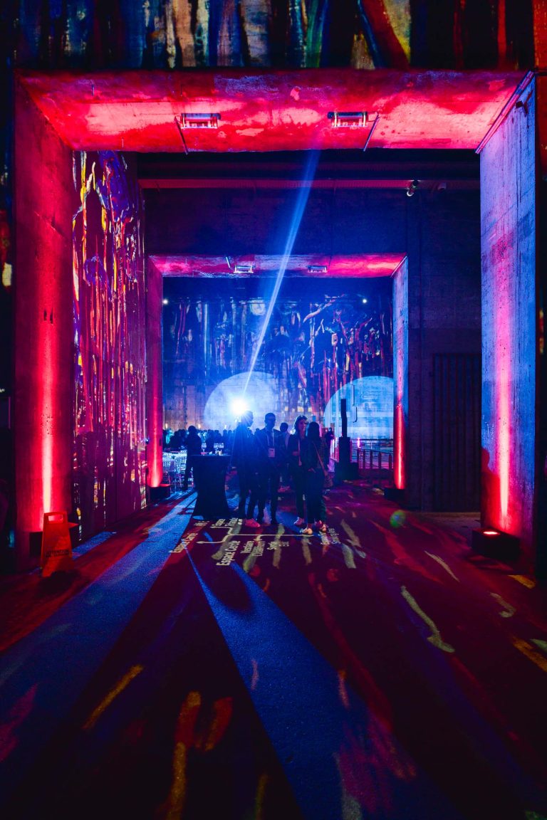 Une exposition d'art dynamique à la French Tech Night 2024 présente des projections colorées et abstraites sur les murs et le sol en béton. La salle est illuminée de lumières rouges, bleues et violettes, créant une atmosphère dynamique. Un groupe de personnes se tient au centre, rétroéclairé par un projecteur blanc brillant, observant l'art qui les entoure.