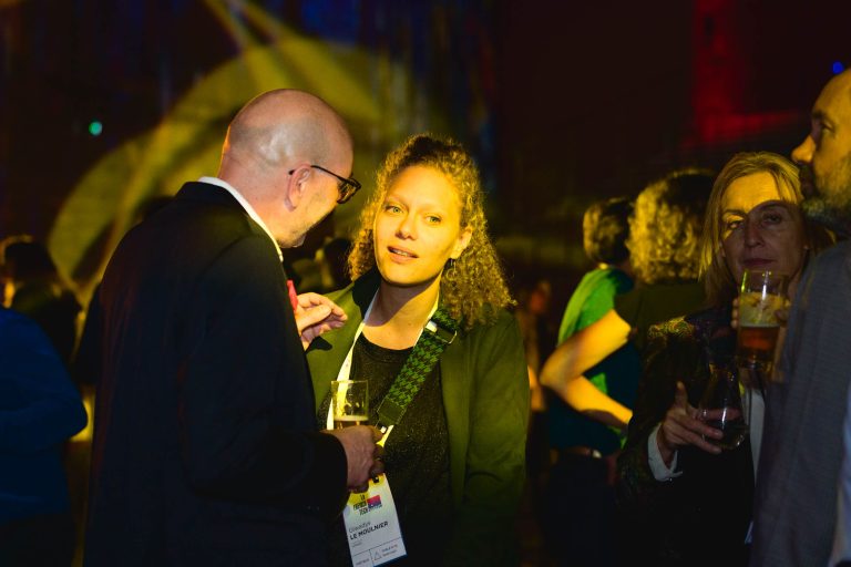 Un groupe de personnes discutent lors de la French Tech Night 2024, un événement avec un éclairage tamisé et des projecteurs colorés. Au premier plan, un homme chauve portant des lunettes et un costume discute avec une femme aux cheveux bouclés tenant un verre. D'autres participants, tenant des verres, sont visibles à l'arrière-plan, éclairés par l'éclairage d'ambiance.