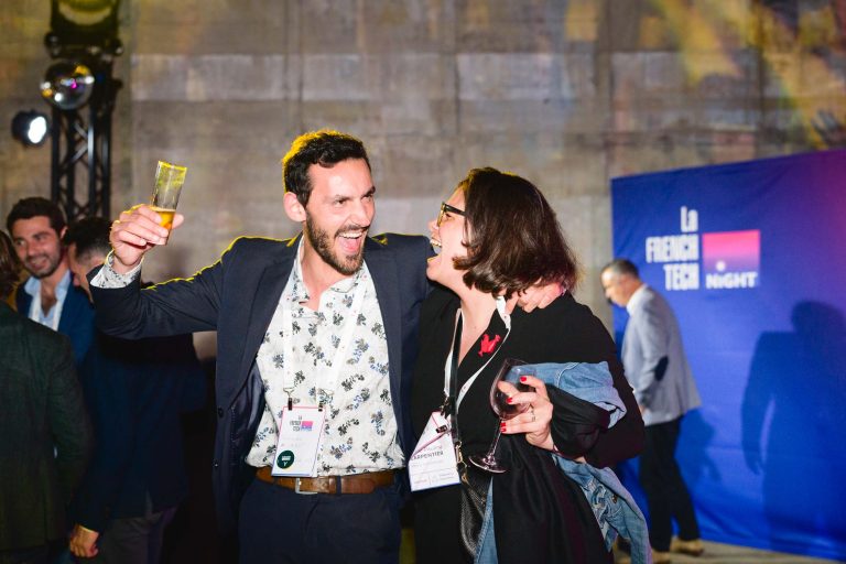 Un homme et une femme interagissent joyeusement lors de l'événement French Tech Night 2024, chacun tenant une boisson. L'homme a un bras levé avec un verre de liquide orange, tandis que la femme tient un verre de vin. Tous deux sourient et portent des badges nominatifs. Une bannière bleue en arrière-plan indique « La French Tech Night ».