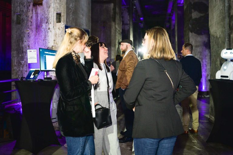 Un groupe de personnes est réuni à la French Tech Night 2024, discutant et souriant. Le décor semble industriel avec des piliers et des murs en béton illuminés par un éclairage violet. Certains tiennent des boissons, tandis qu'un robot est positionné à droite. La tenue varie du décontracté au décontracté professionnel.