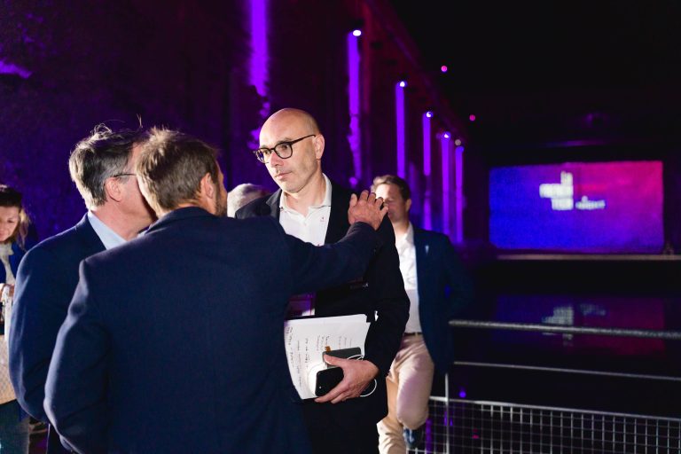 Lors de la French Tech Night 2024, un groupe de personnes discute sous un éclairage violet vif. Un homme portant des lunettes, tenant des papiers et un téléphone, est tapoté sur l'épaule par un autre homme. Un grand écran affiche un logo illisible dans ce lieu moderne et industriel.