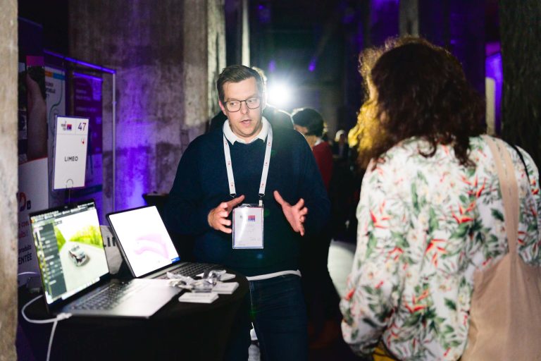 Un homme portant des lunettes fait des gestes tout en expliquant quelque chose à une femme aux cheveux bouclés sur le stand de l'exposition French Tech Night 2024. Le stand dispose de deux ordinateurs portables ouverts affichant des graphiques, et un cordon avec une carte d'identité est suspendu autour du cou de l'homme. La scène est faiblement éclairée avec un éclairage violet en arrière-plan.