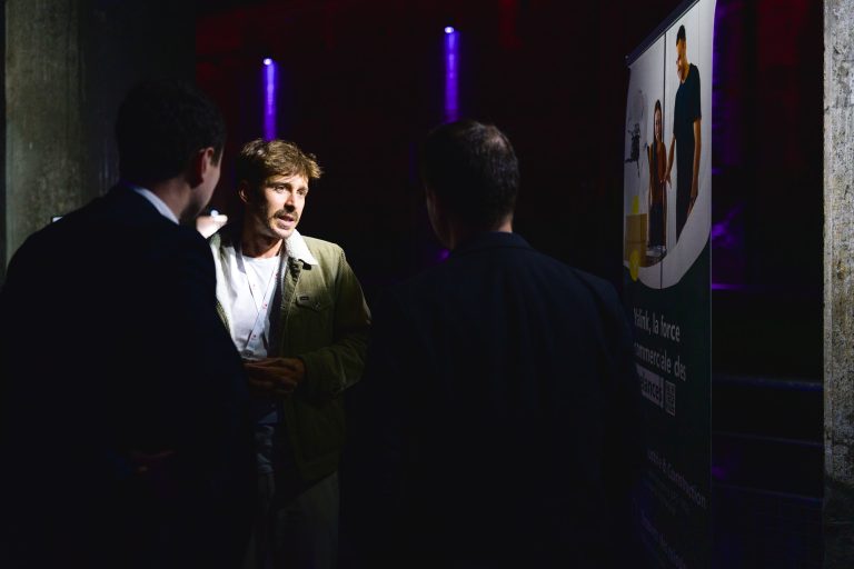 Trois hommes discutent dans un environnement faiblement éclairé lors de la French Tech Night 2024. L'homme au milieu, vêtu d'une veste verte, est bien éclairé et semble parler. Deux hommes, vêtus de costumes sombres, tournent le dos à la caméra. Une bannière partiellement visible se trouve à l'arrière-plan.