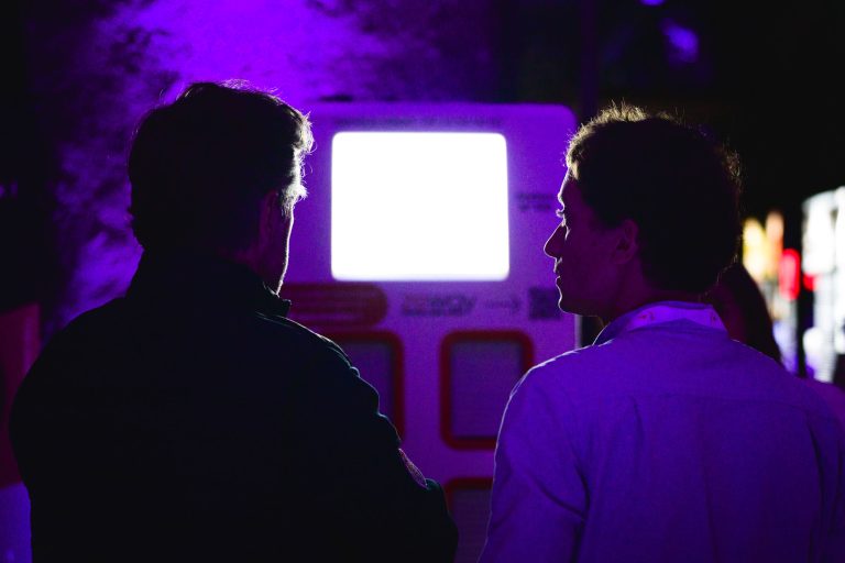 Deux personnes sont photographiées de dos, debout devant un écran très éclairé dans un environnement faiblement éclairé par un éclairage violet. L'une semble porter une veste sombre et l'autre une chemise claire. Elles semblent être engagées dans une conversation tout en observant l'écran lors de la French Tech Night 2024.