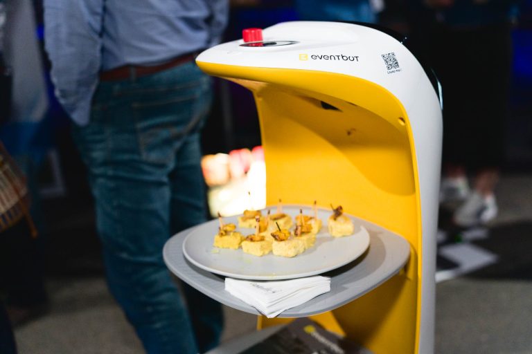 Un robot jaune et blanc nommé « eventbot » sert une assiette de bouchées d'amuse-gueules, chacune avec un cure-dent, sur sa surface plane. Il tient également une petite pile de serviettes. L'arrière-plan montre des personnes debout, indiquant un rassemblement social ou un événement à la French Tech Night 2024.