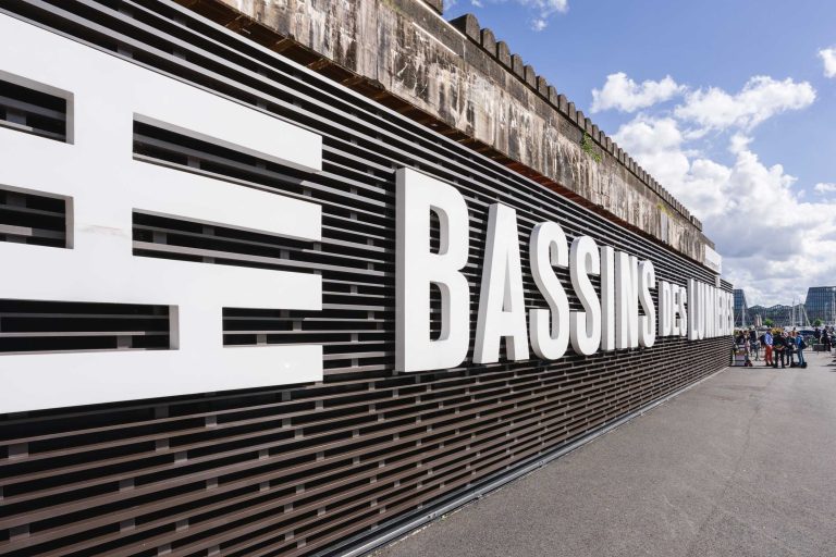 Une grande enseigne sur laquelle est inscrit « BASSINS DES LUMIÈRES » trône sur une façade à claire-voie horizontale et moderne. Le texte blanc contraste avec le fond sombre. Des passants sont visibles à droite de l'enseigne, marchant sur le trottoir. Peut-être se dirigent-ils vers la French Tech Night 2024. Le ciel est partiellement nuageux au-dessus de la structure.