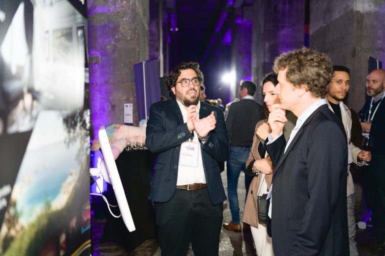 Un homme portant des lunettes et un costume sombre parle et fait des gestes avec animation près d'un écran. Il se tient debout avec un groupe de personnes attentives en tenue professionnelle, dans une salle faiblement éclairée par un éclairage violet. L'atmosphère évoque la French Tech Night 2024, faisant allusion à un événement ou une présentation officielle.