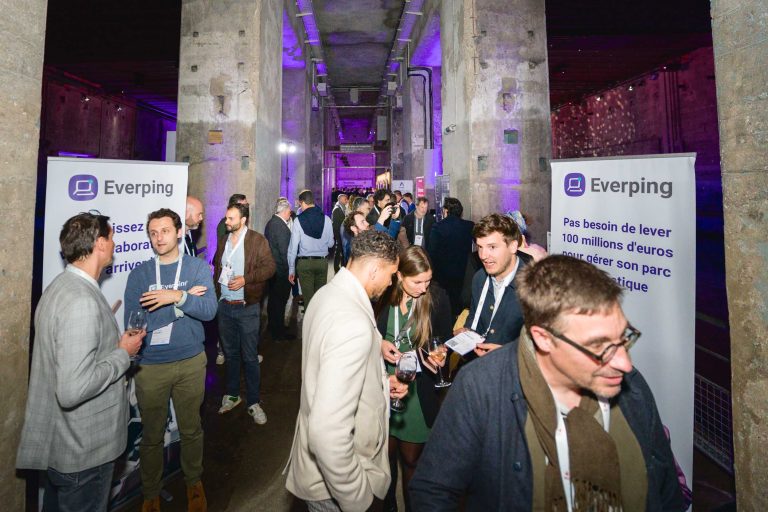 Un grand groupe de personnes à la French Tech Night 2024 dans un lieu de style industriel avec des piliers en béton et un éclairage violet. Deux bannières de chaque côté affichent le logo et le nom « Everping ». Les participants discutent, tiennent des boissons et portent des badges d'événement.