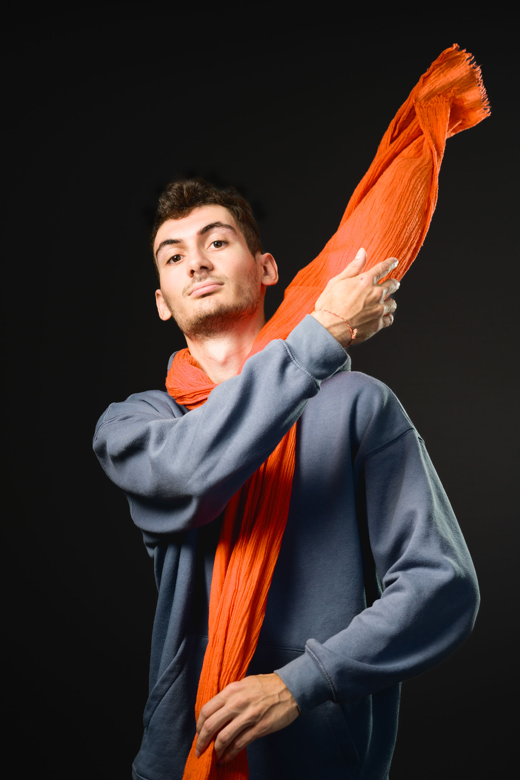 Une personne aux cheveux courts et foncés porte un sweat-shirt bleu et pose avec assurance sur un fond sombre. Elle enroule de manière ludique un foulard orange autour de son cou, avec une extrémité du foulard tenue haute sur son épaule. L'expression de son visage semble déterminée et concentrée, incarnant l'esprit des portraits professionnels à Bordeaux.