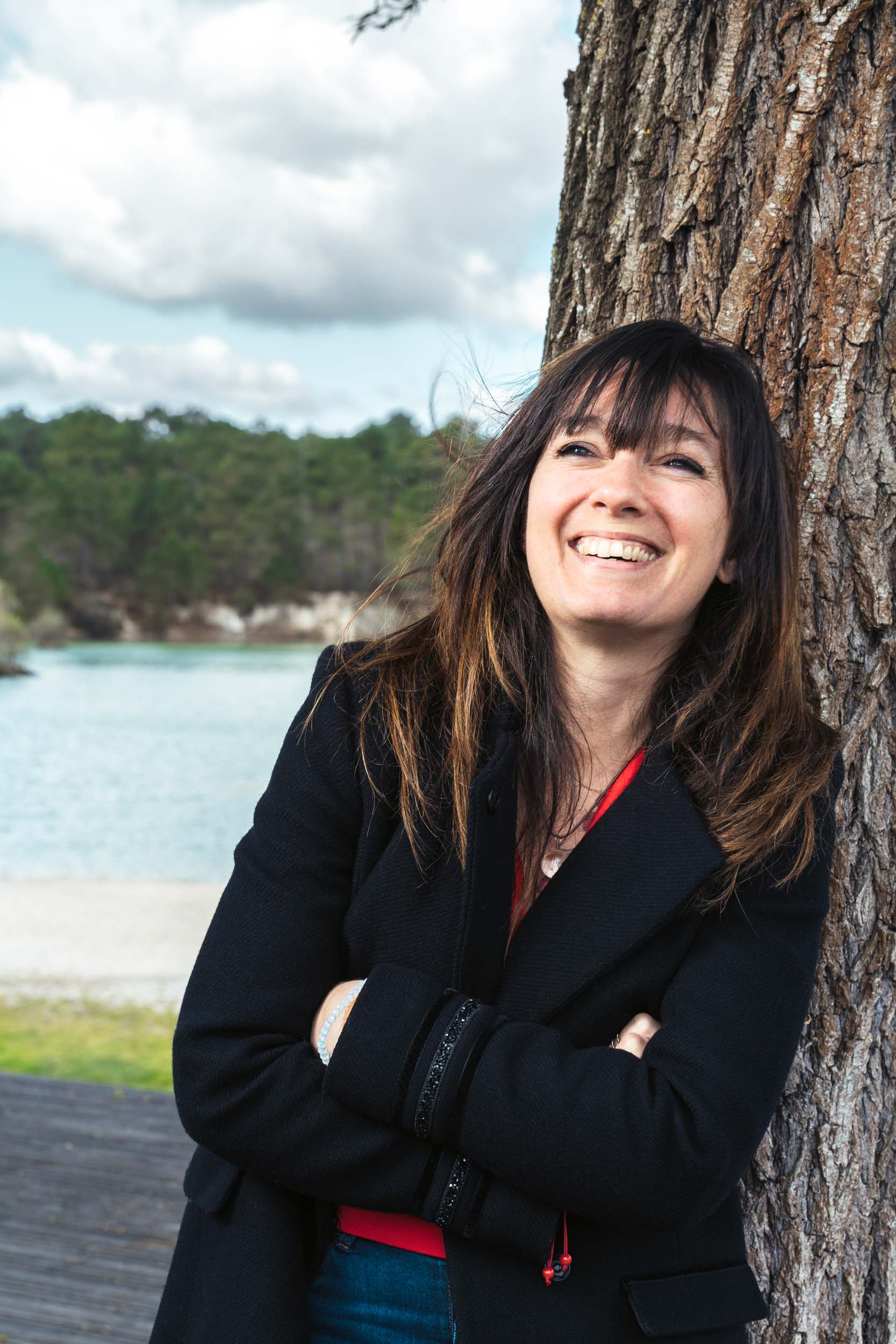 Une femme aux longs cheveux bruns, vêtue d'un manteau noir, s'appuie contre un tronc d'arbre, souriant largement. Elle se tient debout, les bras croisés, devant un paysage pittoresque composé d'un lac, d'arbres verts et d'un ciel nuageux. Son attitude est détendue et joyeuse, parfaite pour les portraits professionnels à Bordeaux.