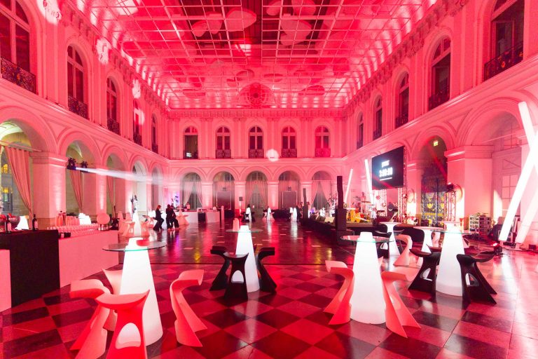 Une grande salle aux hauts plafonds illuminés par un éclairage rouge, décorée pour la Soirée Annuelle du Groupe Betclic. Des tables et des chaises modernes et lumineuses sont disposées sur le sol en damier. De grandes fenêtres cintrées, des colonnes et une mezzanine accentuent l'espace tandis que des personnes en arrière-plan engagent des conversations.