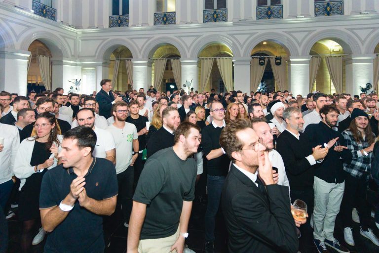 Une grande foule se rassemble à l'intérieur lors de la soirée annuelle du Groupe Betclic. Les participants se tiennent dans une salle spacieuse et bien éclairée, avec de hauts plafonds et des ouvertures en arc sur les murs. De nombreux participants se concentrent sur une scène ou un orateur invisible, semblent engagés et applaudissent. L'atmosphère semble formelle mais animée.