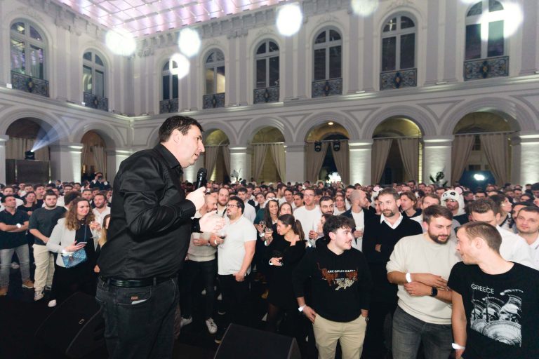 Un homme en chemise noire se tient sur scène, parle au micro et s'adresse à un large public dans une grande salle aux fenêtres cintrées et à l'éclairage ambiant. La foule, vêtue de tenues décontractées pour la Soirée annuelle du Groupe Betclic, fait face à la scène avec attention. La salle est élégante, avec de hauts plafonds et une atmosphère festive.