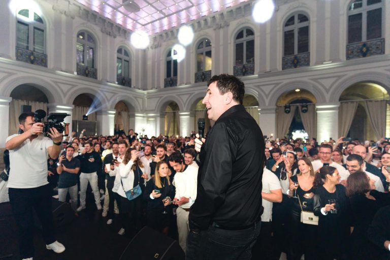 Un homme en chemise noire parle dans un micro, face à un large public lors de la soirée annuelle du groupe Betclic dans une salle décorée. La foule semble engagée, avec beaucoup de sourires, d'applaudissements et de boissons à la main. Un caméraman à gauche capture la scène, mettant en valeur les hauts plafonds et les fenêtres cintrées de la salle.