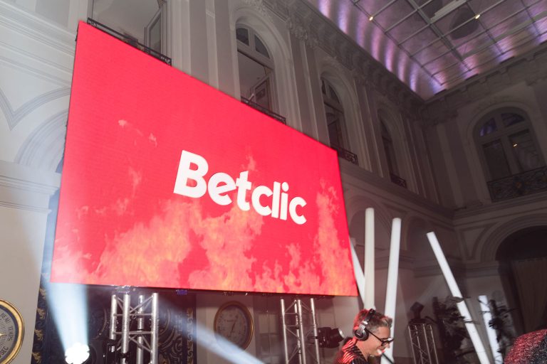 Un grand écran rouge éclairé avec le texte blanc « Betclic » est affiché dans un lieu intérieur aux hauts plafonds et à l'architecture élégante. Sous l'écran se tient une personne portant des écouteurs, peut-être un DJ, avec du matériel d'éclairage et des gens autour. La Soirée annuelle du Groupe Betclic est dotée de fenêtres cintrées et d'un plafond en verre.