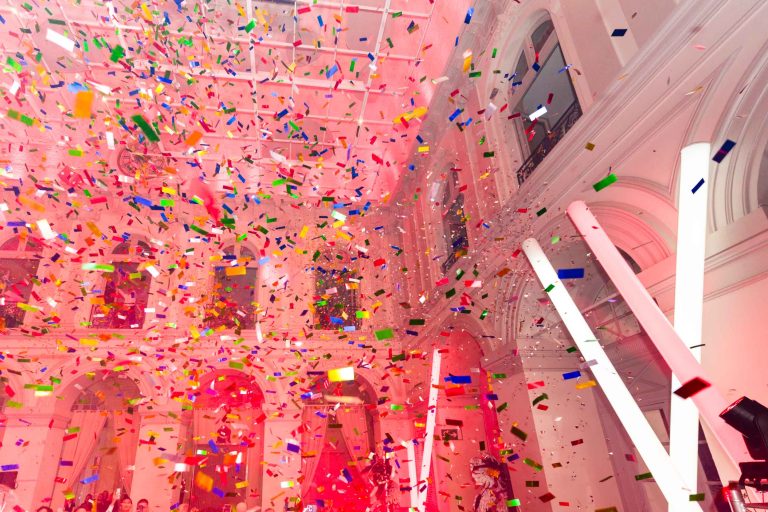 Une grande salle aux hauts plafonds et aux fenêtres cintrées, décorée de confettis colorés tombant du ciel. La salle est baignée d'une lumière rouge et de grands tubes lumineux s'étendent du sol vers le haut. La scène animée et festive suggère l'ambiance joyeuse du Groupe Soirée Annuelle Betclic.