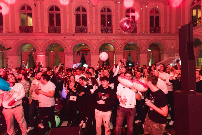 Une foule animée fait la fête dans une grande salle aux hautes arches et au décor richement décoré lors de la Soirée annuelle du Groupe Betclic. Les gens dansent, applaudissent et lancent des confettis. L'éclairage est chaleureux et coloré, ajoutant à l'atmosphère festive. Des ballons et de gros objets gonflables sont dispersés dans toute la salle.