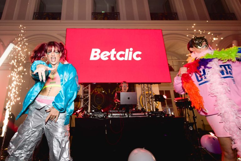 Deux artistes dansent avec énergie devant une cabine de DJ lors d'un événement en salle. À gauche, un danseur porte une veste bleue, un pantalon argenté et des lunettes de soleil. À droite, un autre danseur porte une tenue rose avec un boa en plumes colorées. Derrière eux, un écran affiche le logo « Betclic » lors de la Soirée annuelle du Groupe Betclic.