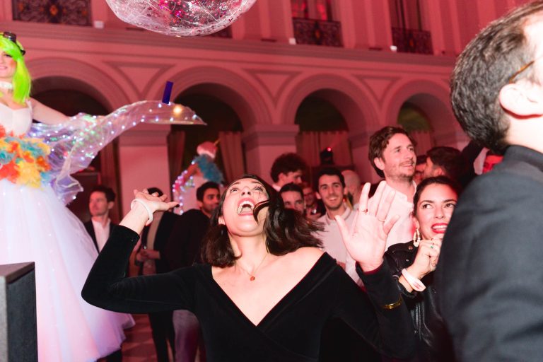 Lors de la soirée annuelle animée du groupe Betclic, les gens dansent et s'amusent. Au premier plan, une femme aux cheveux noirs et à la robe noire sourit largement et lève les mains. D'autres invités, vêtus de tenues diverses, sont à l'arrière-plan. Un artiste en costume coloré avec une perruque verte est sur des échasses.