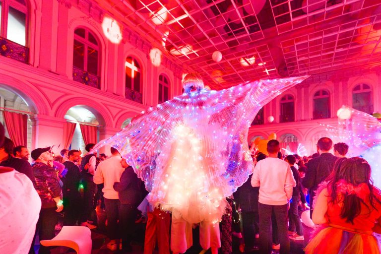 Une personne vêtue d'un costume illuminé avec des ailes d'ange remplies de lumières multicolores se tient au-dessus d'une foule de personnes assistant à la soirée annuelle animée du groupe Betclic, dans un bâtiment décoré avec de hauts plafonds et des arches. La salle rayonne d'une lueur rouge provenant de l'éclairage ambiant.