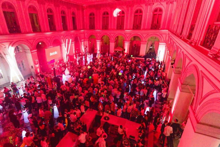 Une foule nombreuse profite d'une soirée annuelle animée organisée par le groupe Betclic dans une grande salle illuminée de lumières rouges. Le lieu est doté de hauts plafonds et de plusieurs arches. Les invités se rassemblent autour des tables, discutent, dansent et regardent une performance sur une scène avec un logo « Reddit » projeté en arrière-plan. Des ballons flottent au-dessus de la foule.