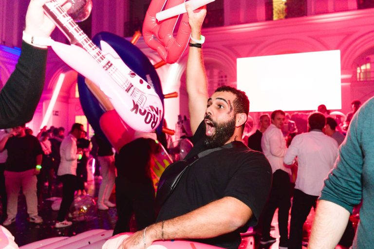 Un homme barbu agite avec enthousiasme une guitare gonflable d'une main tout en levant l'autre main en poing lors de la Soirée annuelle animée du Groupe Betclic. L'arrière-plan est rempli de gens, d'éclairages colorés et de décorations élaborées, créant une atmosphère festive.