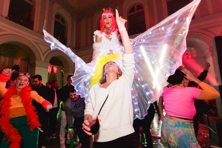 Une soirée annuelle animée du Groupe Betclic dans une grande salle aux lumières colorées. Un artiste aux cheveux roses et aux ailes illuminées s'élève au-dessus de la foule. Les participants, vêtus de tenues et d'accessoires colorés, dansent avec énergie autour d'eux. L'ambiance est festive et dynamique, remplie de mouvement et de joie.