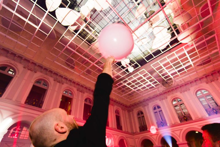 Un personnage au crâne chauve et à la barbe, vêtu de vêtements sombres, tend la main vers un gros ballon rose. La soirée annuelle du Groupe Betclic se déroule dans une salle élégante avec des fenêtres cintrées, des lumières claires et un plafond en forme de grille. L'éclairage est à dominante rose et rouge, créant une atmosphère festive.