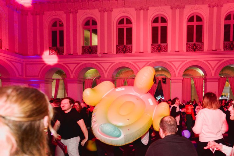 Des gens se rassemblent dans une grande salle richement décorée avec de hautes fenêtres cintrées et des balcons pour la Soirée annuelle du Groupe Betclic. Une personne dans la foule porte un grand flotteur de piscine gonflable coloré. La salle est éclairée de lumières roses et rouges, créant une atmosphère festive.