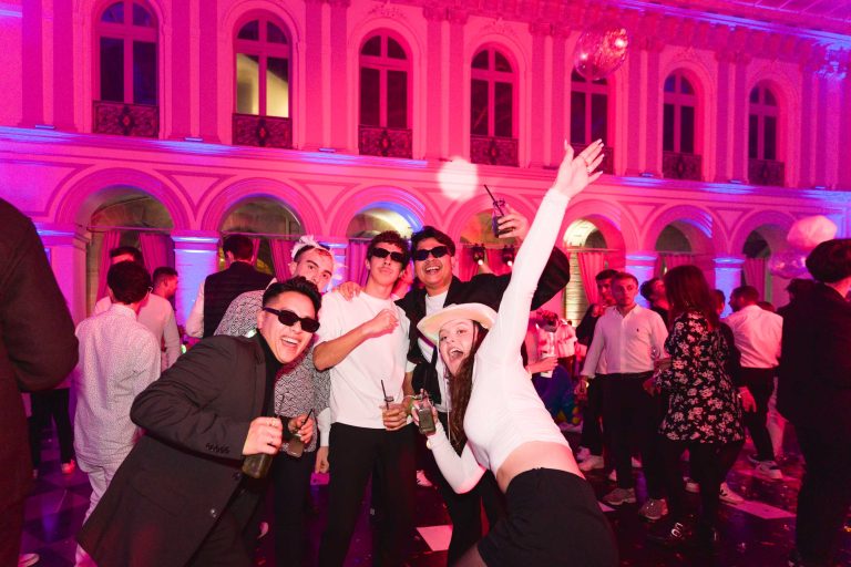 Un groupe animé de cinq personnes est habillé en tenue de soirée, danse et pose pour la caméra lors de la Soirée Annuelle du Groupe Betclic, illuminée par un éclairage rose et violet vibrant. Dans une grande salle aux fenêtres cintrées et à l'architecture ornementée, certains portent des lunettes de soleil tandis que l'un d'eux agite énergiquement son bras.