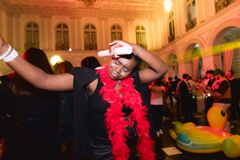 Une personne dans un boa de plumes rouge vif danse joyeusement lors de la Soirée annuelle du groupe Betclic. Elle tient un morceau de papier enroulé, incline la tête vers l'arrière et ferme les yeux. L'arrière-plan présente des fêtards et des jouets de piscine gonflables dans une salle spacieuse, à haut plafond et dotée de hautes fenêtres cintrées.