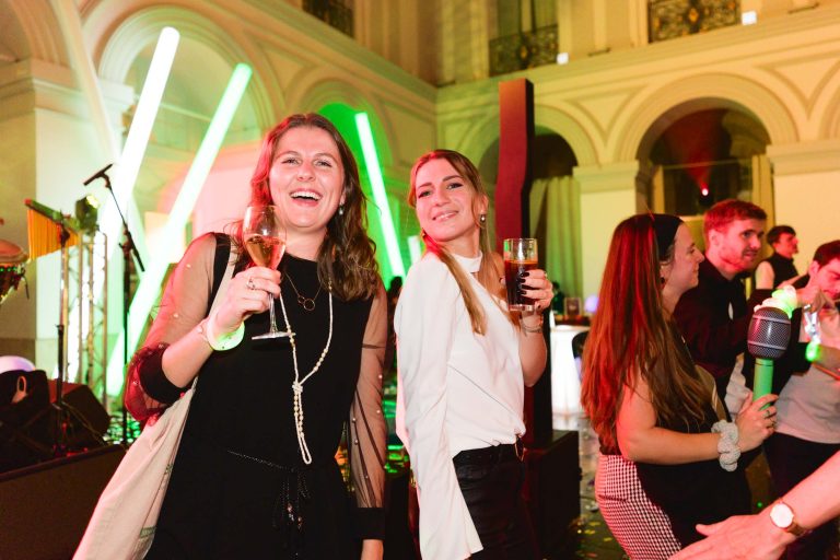 Deux femmes se tiennent debout et sourient lors d'un événement intérieur animé avec des lumières vertes au néon et une arche en arrière-plan. La femme de gauche tient un verre de champagne et celle de droite une bière. Toutes deux sont habillées avec élégance et apprécient la Soirée annuelle du groupe Betclic. D'autres personnes à l'arrière-plan discutent et dansent.