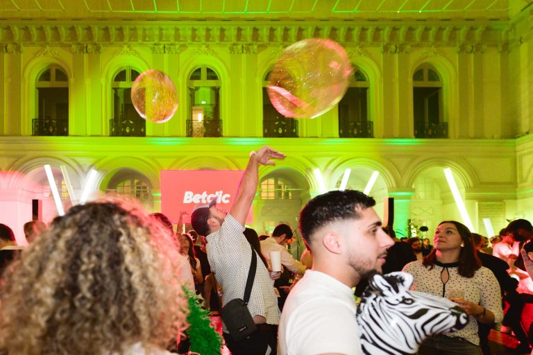 Une soirée annuelle animée en intérieur par le groupe Betclic avec des lumières colorées illuminant une salle élégante aux hautes arches. Les invités dansent et interagissent, avec une personne lançant une grosse balle gonflable translucide. L'ambiance est festive, avec des personnes tenant des boissons, portant des tenues décontractées et une personne tenant une peluche zèbre.