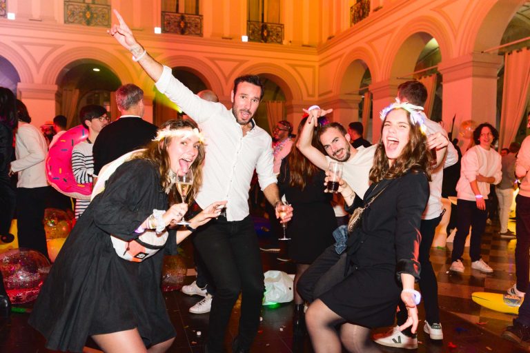 Un groupe de personnes animées dansent lors de la Soirée annuelle festive du groupe Betclic avec de hauts plafonds et des fenêtres cintrées. Deux femmes et un homme au premier plan posent avec énergie, tenant des boissons et portant des accessoires lumineux. La salle est remplie d'autres participants qui se mêlent et profitent de la célébration.