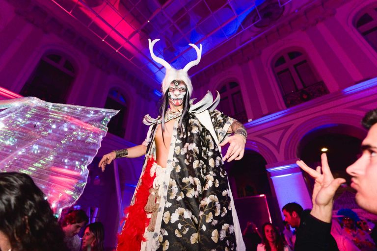 Un artiste vêtu d'un costume élaboré avec des bois de cerf, de la peinture pour le visage et des accessoires en forme d'ailes divertit la foule lors de la Soirée annuelle du Groupe Betclic, une soirée vibrante et colorée. L'arrière-plan présente un bâtiment aux détails architecturaux avec des fenêtres cintrées illuminées par des lumières roses et bleues, ajoutant à l'atmosphère festive.