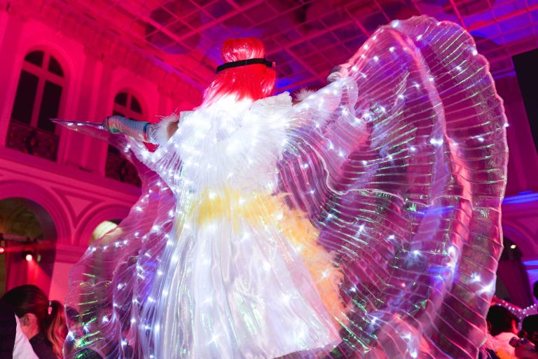 Lors de la Soirée annuelle du Groupe Betclic, une artiste aux cheveux roux fluo et à la tenue blanche étincelante déploie de grandes ailes illuminées par des lumières. En arrière-plan, un élégant bâtiment aux fenêtres cintrées est illuminé de rose et de violet, créant une atmosphère vibrante et magique. Des spectateurs sont partiellement visibles en bas.