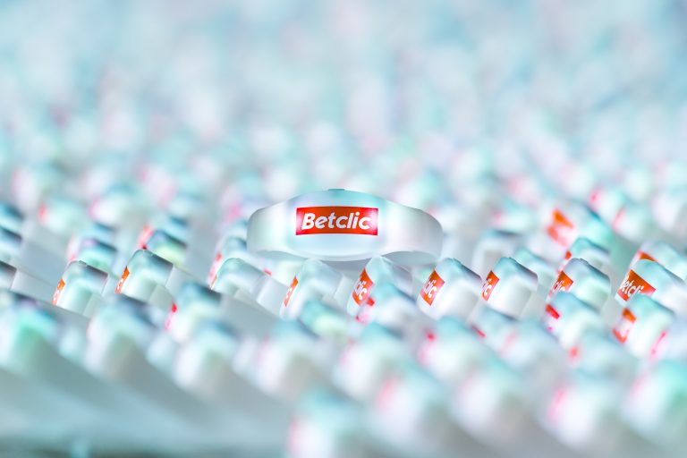 Gros plan sur plusieurs bracelets blancs avec le logo « Betclic » en rouge, rappelant la Soirée annuelle du Groupe Betclic. Les bracelets sont disposés selon un motif semi-uniforme, créant une texture et une profondeur visuellement attrayantes. L'accent est mis sur le bracelet central, tandis que les autres se fondent dans l'arrière-plan.