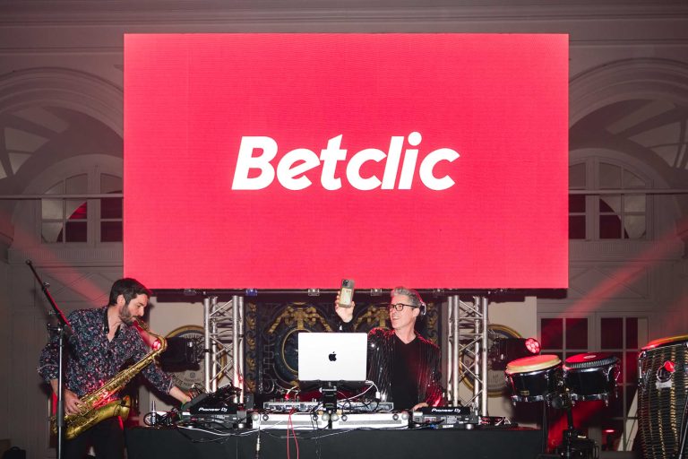 Deux musiciens se produisent sur scène sous un grand écran rouge affichant le mot « Betclic » lors de la Soirée Annuelle du Groupe Betclic. À gauche, un saxophoniste joue, tandis qu'à droite, un DJ se tient debout devant une console de mixage, une bouteille à la main et un casque sur le visage, sur un décor grandiose de détails architecturaux complexes et d'éclairages vibrants.