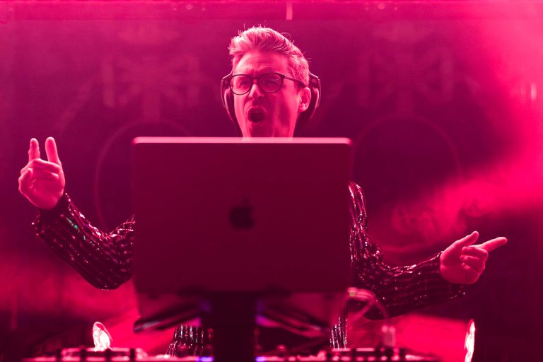 Un DJ aux cheveux blonds et aux lunettes se tient derrière un ordinateur portable sur un support, et joue avec énergie lors de la Soirée annuelle du Groupe Betclic. Il porte des écouteurs et une veste scintillante, entouré d'un éclairage de scène rose et violet, créant une atmosphère dynamique. Sa bouche est ouverte, comme s'il chantait ou criait dans la foule.