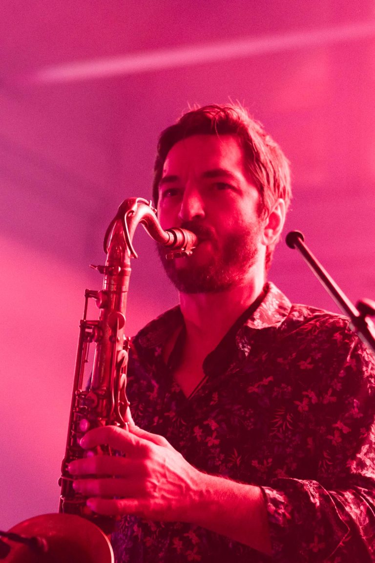 Un homme barbu joue du saxophone sous un éclairage rouge vif lors de la Soirée annuelle du Groupe Betclic. Il porte une chemise boutonnée à motifs et se tient devant un arrière-plan flou sur lequel on peut voir un pied de micro. La lumière ambiante projette une teinte rougeâtre distincte, évoquant un environnement musical vivant et atmosphérique.