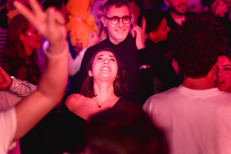 Une femme en haut noir, souriante et regardant vers le haut, est entourée de gens à la Soirée annuelle du Groupe Betclic dans un décor de fête peu éclairé et animé. La scène est baignée d'un éclairage rose et violet. Un homme avec des lunettes et un chandail foncé est derrière elle, souriant également. Les bras et les mains sont levés en signe de célébration autour d'eux.