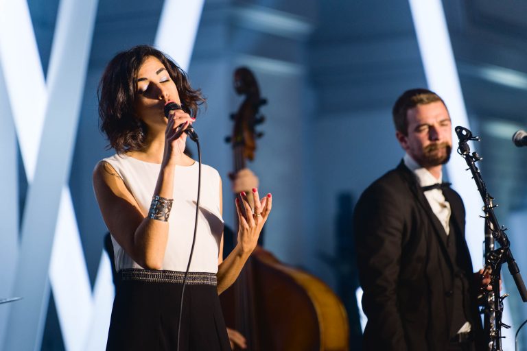 Lors de la Soirée annuelle du Groupe Betclic, une femme en robe blanche et noire chante dans un micro, les yeux fermés et gesticulant de la main gauche. Un homme en costume noir et chemise blanche joue du saxophone à ses côtés. Une contrebasse est visible en arrière-plan, et le décor est éclairé par des lumières blanches en diagonale.